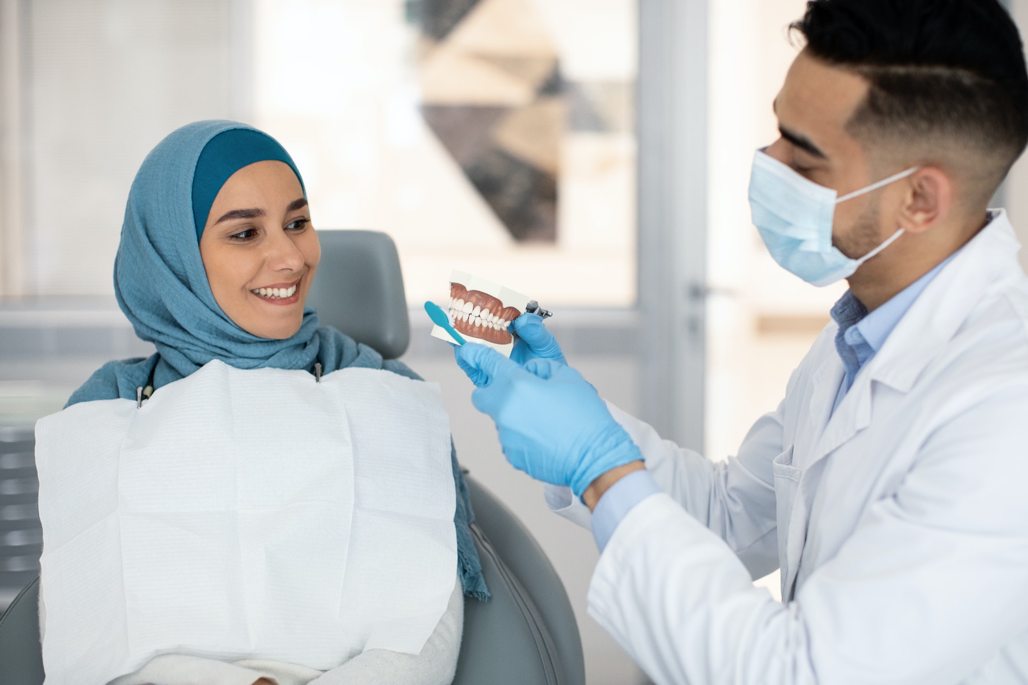 Male dentist holding jaw model, explaining dental hygiene to muslim female patient