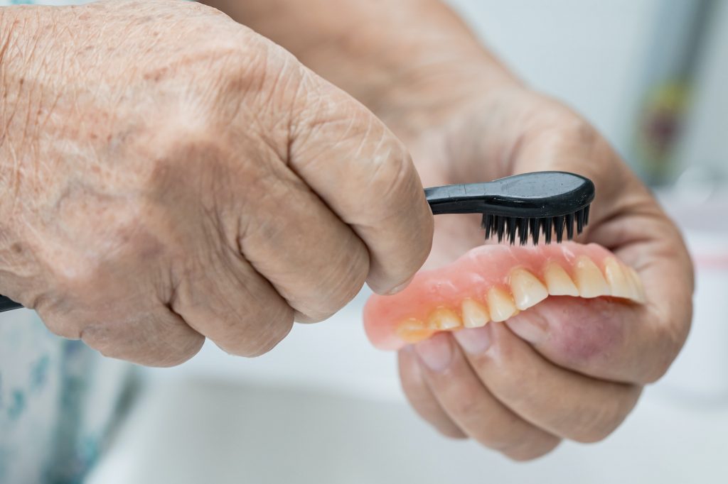 Asian senior woman patient use toothbrush to clean partial denture of replacement teeth.