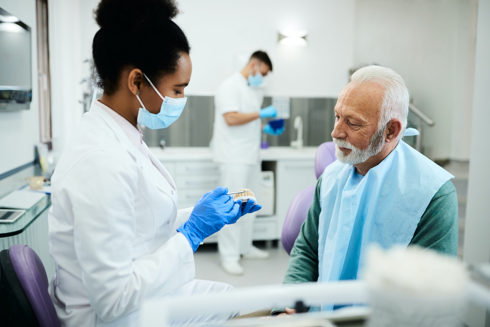 Black dentist using artificial dentures while communicating with senior patient at dentist's office.