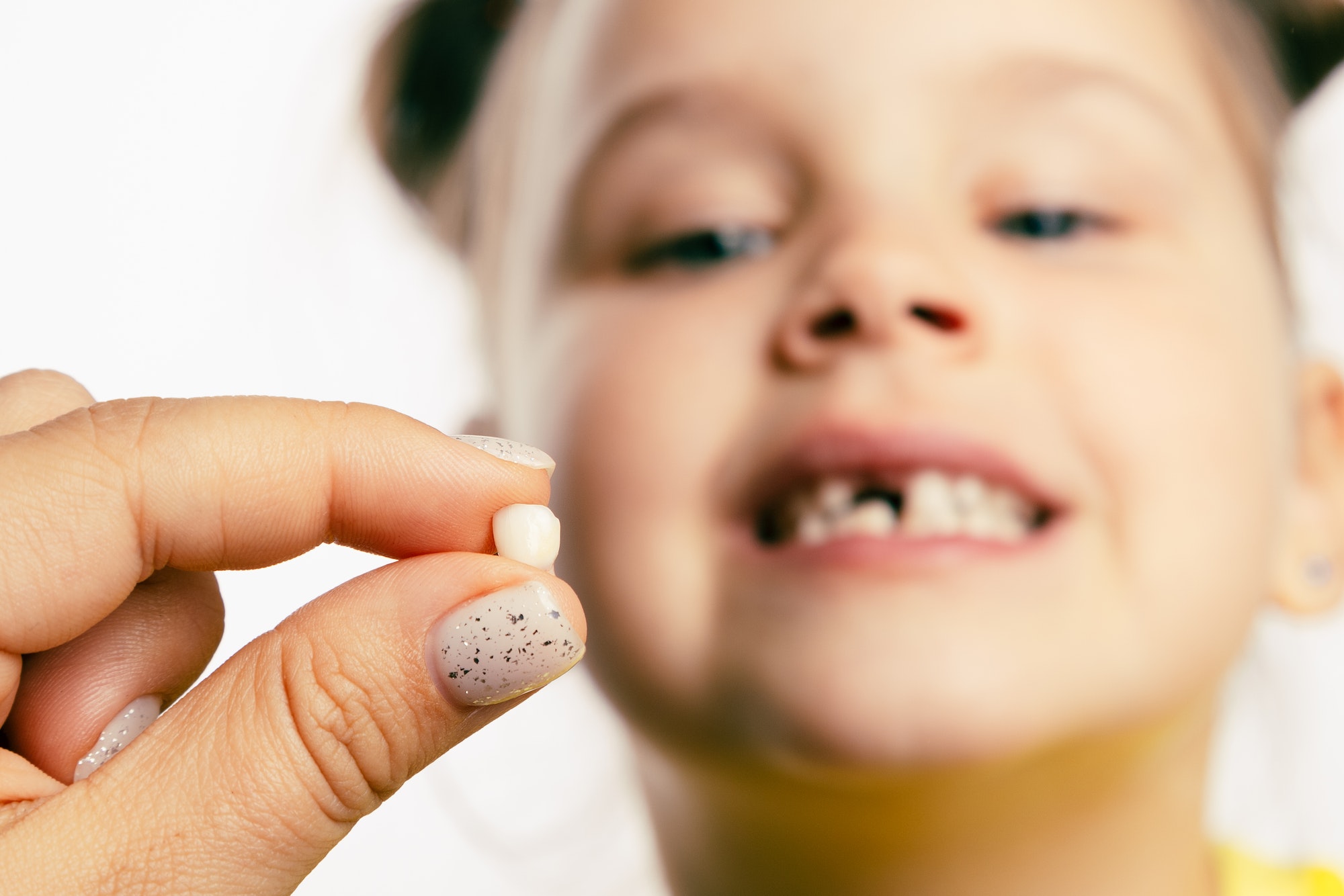 Fingers holding torn out baby milk tooth with little blurred girl face looking at tooth and showing